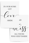 Set of two minimalist wall art prints with the phrases 'In Our Home Let Love Abide' and 'Bless All Those Who Step Inside' in black script and serif fonts, framed in white and styled on a light wood floor
