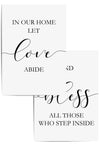 Set of two minimalist wall art prints with the phrases 'In Our Home Let Love Abide' and 'Bless All Those Who Step Inside' in black script and serif fonts, framed in white and styled on a light wood floor