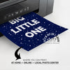 A dark navy blue custom print with white text reading 'BIG LITTLE ONE' and white speckled dots, shown emerging from a printer. promotional text at the bottom about printing at home, online, or locally.