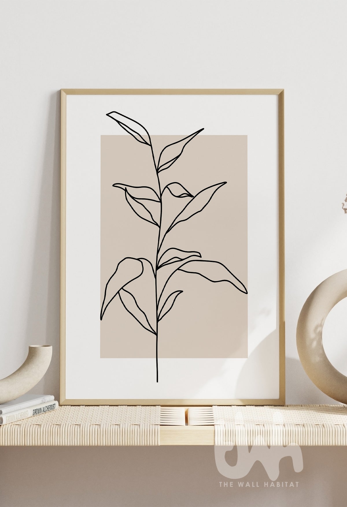 Minimalist line art print of a leafy plant stem, illustrated in black over a warm beige rectangular background, styled in a modern interior setting.