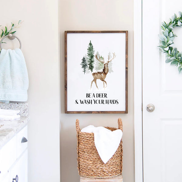Watercolor deer standing in front of evergreen forest with the text “Be a Deer & Wash Your Hands,” rustic woodland bathroom wall art print.