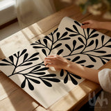 Bold abstract black leaf print on a beige background, shown unframed and being styled on a wooden table in natural daylight.