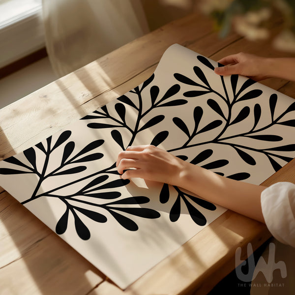 Bold abstract black leaf print on a beige background, shown unframed and being styled on a wooden table in natural daylight.
