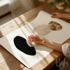 Person holding abstract art print with black and gold circular designs on a wooden table.