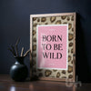 Framed print with 'Born to Be Wild' text on a pink background, leaning against a dark wall.