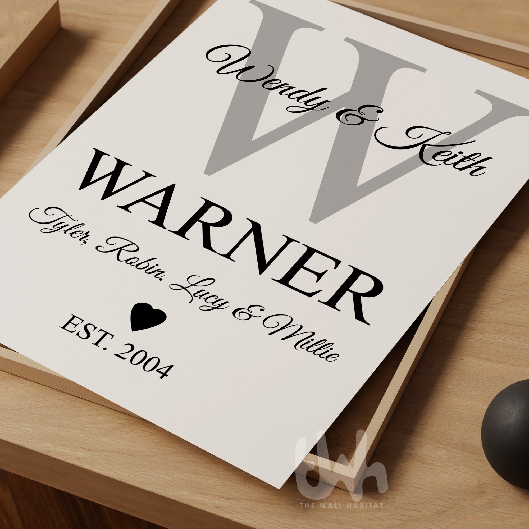 Flat-lay of a custom family monogram print with grey letter W and black personalized text for parents, children, and ‘EST. 2004’, styled on a wooden tray
