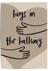 Neutral-toned wall art print leaning against a wooden door in a cozy hallway, featuring minimalist line art of arms and the phrase 'Hugs in the Hallway' over a heart shape