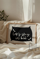 Cozy bedroom scene featuring framed wall art above a bed with the phrase 'Let’s Stay in Bed' inside a black pillow silhouette on a neutral background