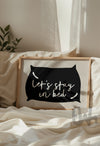 Cozy bedroom scene featuring framed wall art above a bed with the phrase 'Let’s Stay in Bed' inside a black pillow silhouette on a neutral background