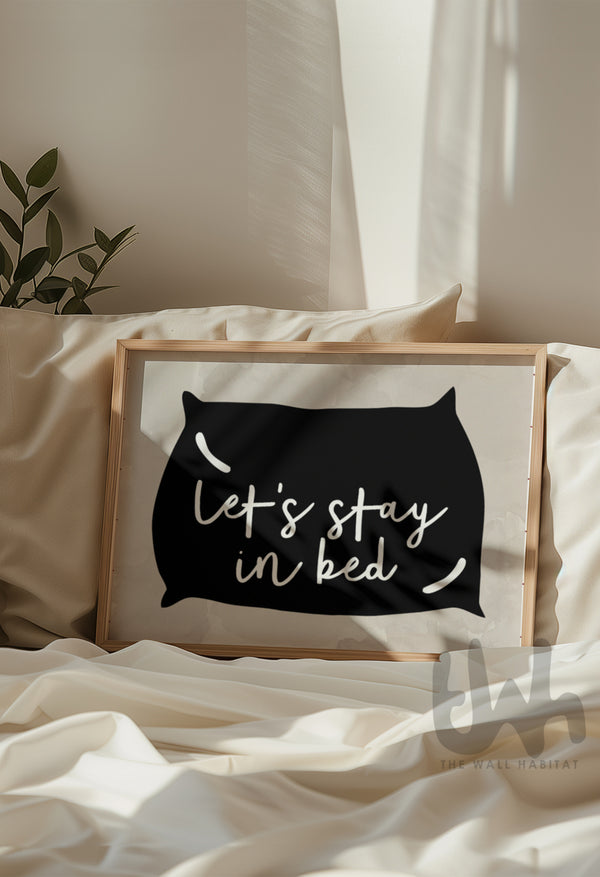 Cozy bedroom scene featuring framed wall art above a bed with the phrase 'Let’s Stay in Bed' inside a black pillow silhouette on a neutral background
