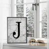 Framed monogram wall art print with a bold black letter 'J' on a grey marble background, styled on a windowsill with a snowy view outside