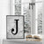 Framed monogram wall art print with a bold black letter 'J' on a grey marble background, styled on a windowsill with a snowy view outside