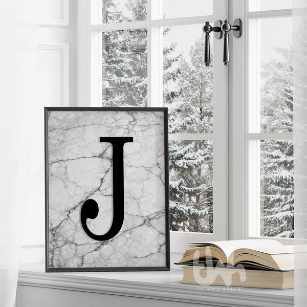Framed monogram wall art print with a bold black letter 'J' on a grey marble background, styled on a windowsill with a snowy view outside