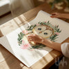 Hands unrolling a sage green owl nursery wall art print with a pink floral wreath and watercolor baby owl – perfect for baby girl woodland décor