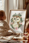 Toddler sitting by a window looking at a framed sage green owl nursery print surrounded by soft pink flowers – baby room styling inspiration