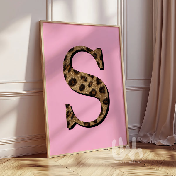 Framed poster with a leopard print letter 'S' on a pink background, placed in a room with light-colored walls and curtains.