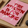 Pink poster with red text 'This kitchen is for dancing' on a wooden surface