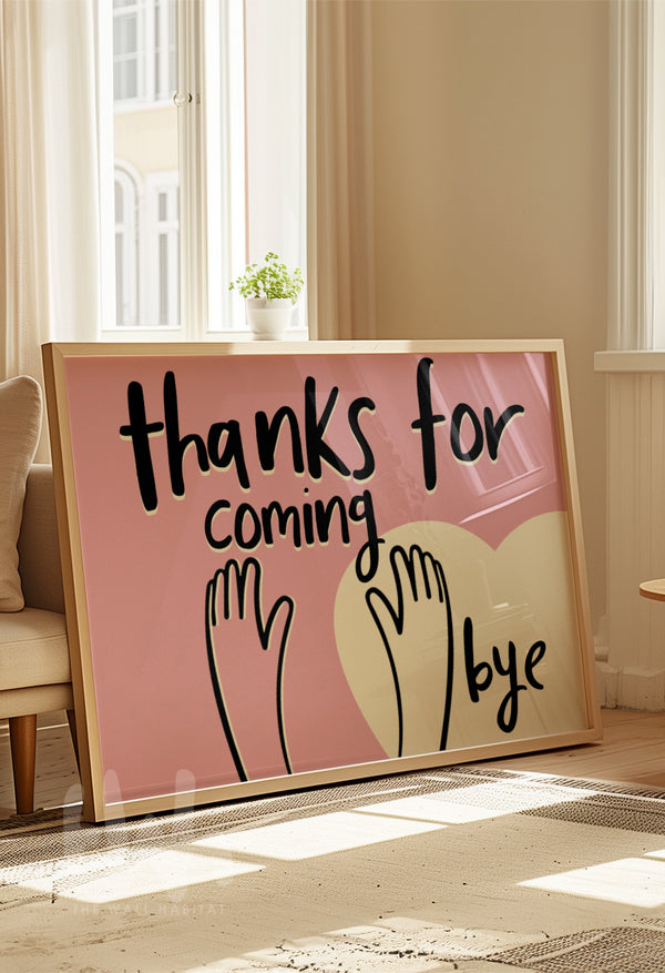 Framed pink wall art print with black handwritten text reading 'Thanks for coming, bye' and hand illustrations over a heart, leaning against the wall in a bright living room