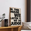 Framed poster with text 'In this house we listen & we do judge' on a wall in a room.