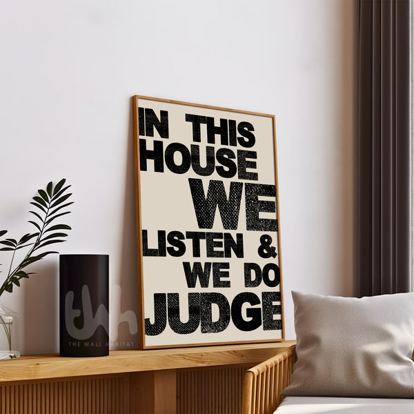 Framed poster with text 'In this house we listen & we do judge' on a wall in a room.