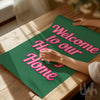 Bold wall art print with pink retro-style text reading 'Welcome to Our Happy Home' on a green background – perfect for cheerful home décor