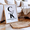 Framed personalized couple’s monogram wall art leaning against a beige sofa, showing initials C and K with a botanical ampersand, names Charlie & Kelly, and wedding date
