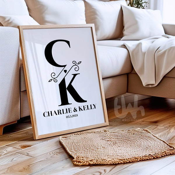 Framed personalized couple’s monogram wall art leaning against a beige sofa, showing initials C and K with a botanical ampersand, names Charlie & Kelly, and wedding date
