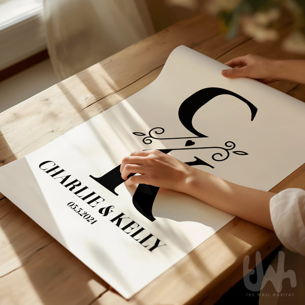 Hands unrolling a personalized wall art print with black initials 'C & K', floral accent, and the names 'Charlie & Kelly' with the date 05.2024 on white background