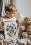 Young child holding a framed watercolor zebra print with pink flowers and sage green foliage, styled in a cozy nursery setting with a teddy bear