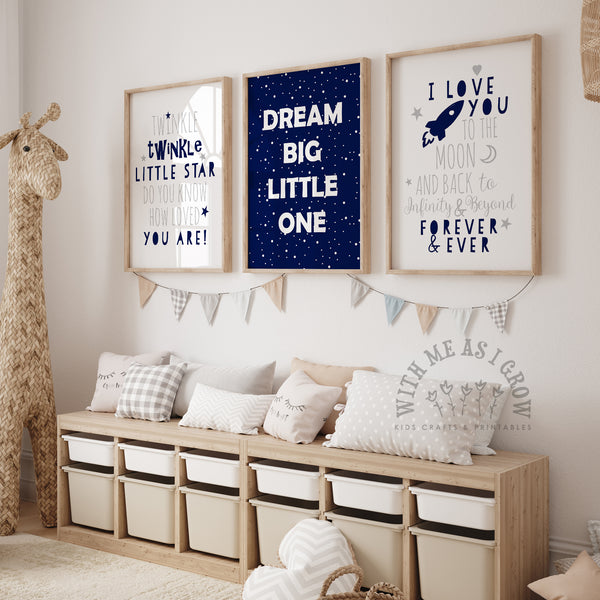 Three framed nursery wall art prints hang above a wooden storage bench in a styled children's room. The center print is navy blue with white text reading 'DREAM BIG LITTLE ONE,' while the left and right prints are white with navy text reading 'I WANTED A TWINKLE LITTLE STAR' and 'I LOVE YOU TO THE MOON AND BACK FOREVER & EVER.