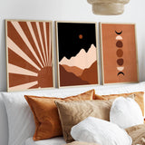 Three framed abstract wall art prints in warm earth tones hang above a bed with white and tan pillows. The left print shows a sunburst, the center shows layered mountains with a sun, and the right shows stacked moon phases.