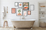 A styled bathroom interior with a freestanding clawfoot tub, white shiplap wall, and a gallery wall of framed botanical and typography art prints. A towel hangs on a wooden hook, a small stool sits beside it, and a shelving unit with folded towels and toiletries is on the right.