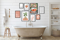 A styled bathroom interior with a freestanding clawfoot tub, white shiplap wall, and a gallery wall of framed botanical and typography art prints. A towel hangs on a wooden hook, a small stool sits beside it, and a shelving unit with folded towels and toiletries is on the right.