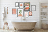 A styled bathroom interior with a freestanding clawfoot tub, white shiplap wall, and a gallery wall of framed botanical and typography art prints. A towel hangs on a wooden hook, a small stool sits beside it, and a shelving unit with folded towels and toiletries is on the right.