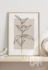 Minimalist line art print of a leafy plant stem, illustrated in black over a warm beige rectangular background, styled in a modern interior setting.