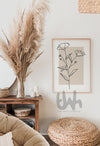 Minimalist line art of blooming flowers with buds, drawn in black on a beige background, framed and styled in a boho living room with natural wood and pampas decor.