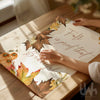 Person holding a decorative paper with autumn-themed design and text on a wooden surface.