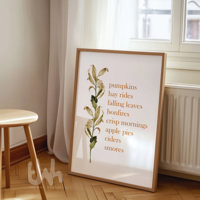 Fall-themed wall art print with a list of autumn favorites like “pumpkins, hay rides, crisp mornings” alongside a watercolor botanical stem, styled in a light wood frame.