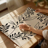Person holding a poster with black leaf pattern on a wooden table.