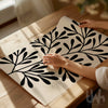 Bold abstract black leaf print on a beige background, shown unframed and being styled on a wooden table in natural daylight.