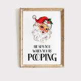 Funny Santa Christmas toilet wall art displayed in a natural oak frame with festive greenery and silver decorations
