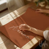 burnt orange print with 'hello Autumn' text on a wooden table, held by a person.