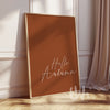 Minimalist burnt orange wall art print with white handwritten script reading “Hello Autumn,” displayed in a wood frame on a fall-styled wall.