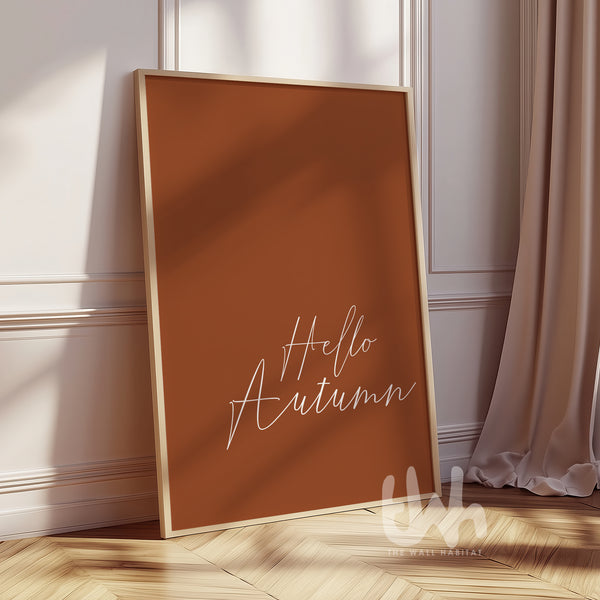 Minimalist burnt orange wall art print with white handwritten script reading “Hello Autumn,” displayed in a wood frame on a fall-styled wall.