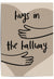 Neutral-toned wall art print leaning against a wooden door in a cozy hallway, featuring minimalist line art of arms and the phrase 'Hugs in the Hallway' over a heart shape