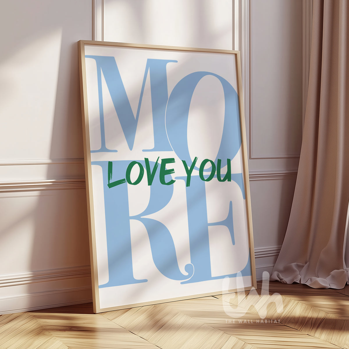 Modern wall art print with the phrase “LOVE YOU MORE” in bold green and baby blue typography, framed and leaning against a white wall in a sunlit room.
