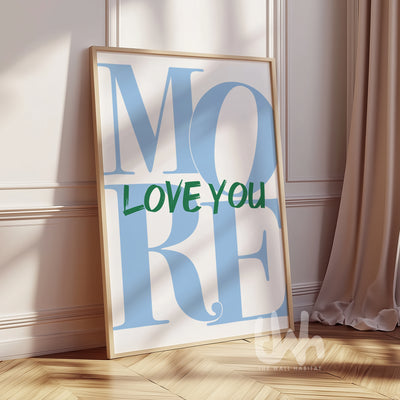 Modern wall art print with the phrase “LOVE YOU MORE” in bold green and baby blue typography, framed and leaning against a white wall in a sunlit room.