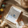 A white framed baby shower print featuring blue elephants, balloons, and bunting with the text 'Congratulations on the arrival of your little elephant!', styled in a warm, sunlit flat-lay with a baby's hand touching the frame. The scene includes a woven placemat, a teddy bear, and baby toys.
