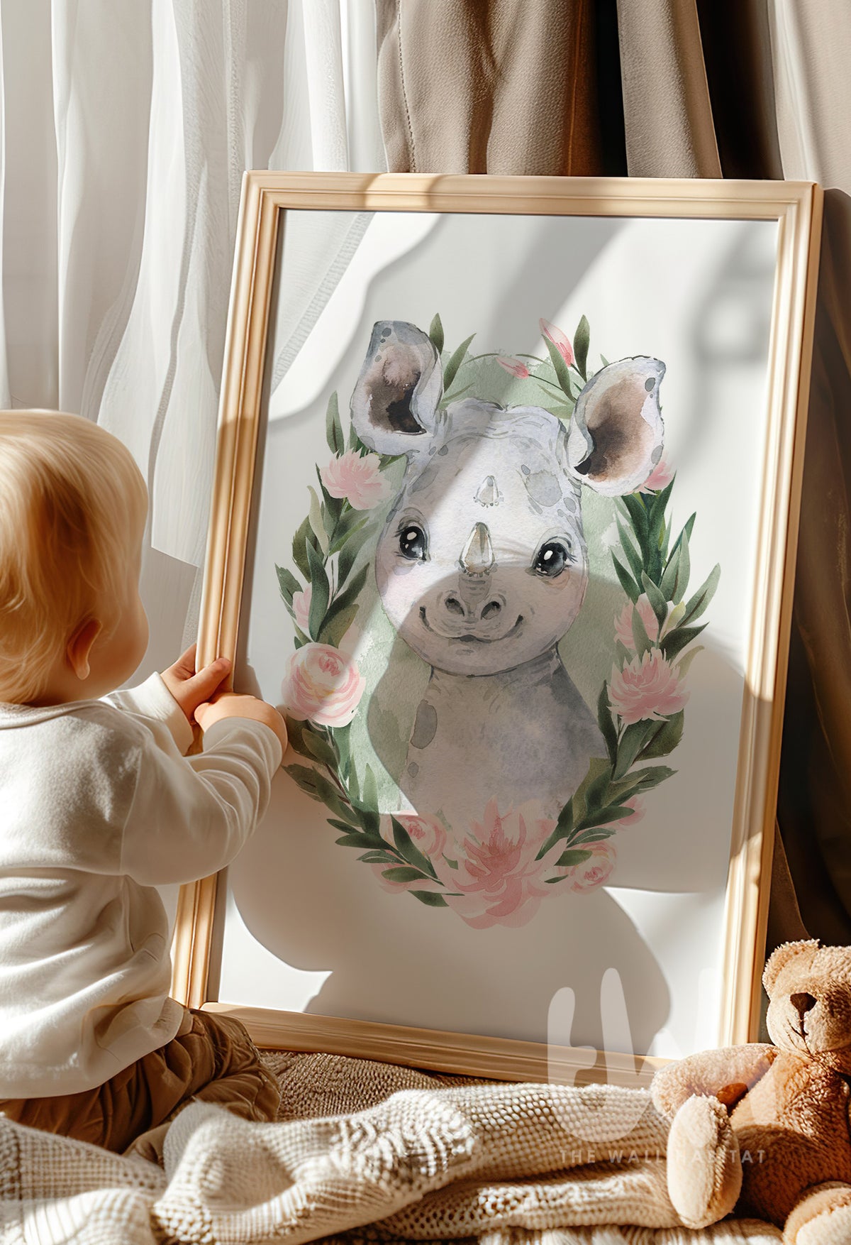 Toddler looking at a framed rhino wall print with pink and green watercolor design – nursery décor styled with a teddy bear