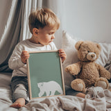 young boy holding a framed green polar bear print while sitting beside a teddy bear on a bed